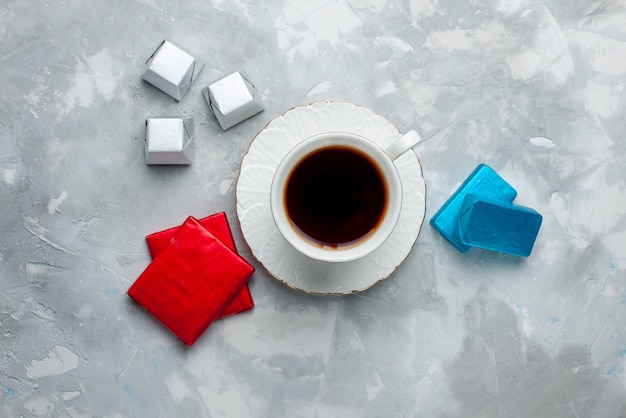 Top view of cup of tea hot inside white cup on glass plate with silver and colored package chocolate candies on light desk, tea drink sweet cookie teatime