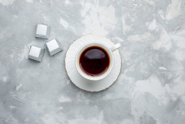 Top view of cup of tea hot inside white cup on glass plate with silver package candies on light desk, tea drink sweet chocolate cookie