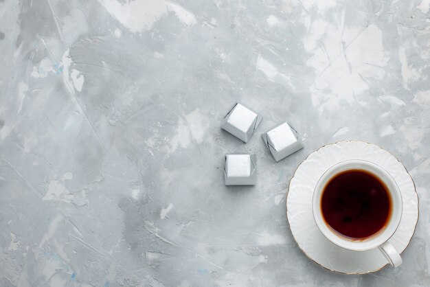 Вид сверху на чашку горячего чая внутри белой чашки на стеклянной тарелке с серебряным пакетом шоколадных конфет на светлом столе, пить сладкое шоколадное печенье
