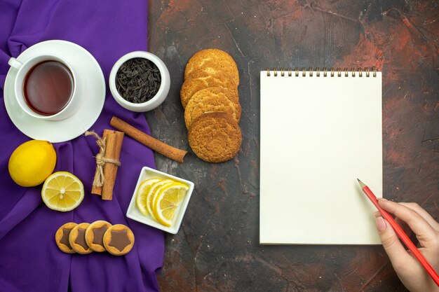 Top view cup of tea lemon slices in bowl cinnamon sticks tea in bowl on purple tablecloth notepad on dark red table