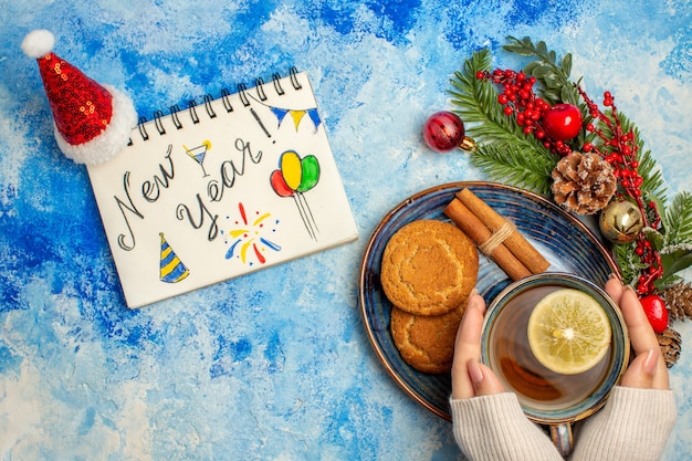 Top view cup of tea lemon slices cinnamon sticks biscuits in saucer new year written on notepad santa hat on blue table