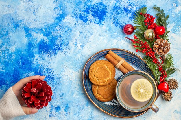 Top view cup of tea lemon slices cinnamon sticks biscuits on saucer red pinecone in female hand on blue table free place