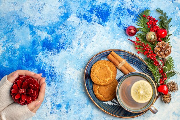 Top view cup of tea lemon slices cinnamon sticks biscuits on saucer red pinecone in female hand on blue table free space