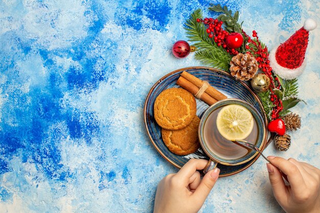 Top view cup of tea lemon slices cinnamon sticks biscuits in saucer xmas tree branch on blue table free space