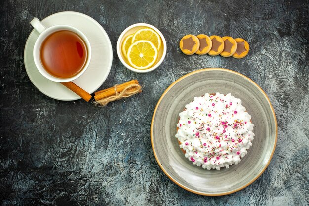 Top view cup of tea lemon slices in small saucer cake on plate biscuits cinnamon sticks on dark table