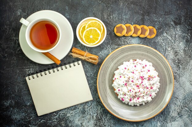 Top view cup of tea lemon slices in small saucer cake on plate biscuits cinnamon sticks notebook on dark table