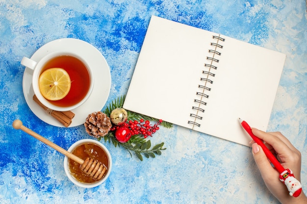 Vista dall'alto tazza di penna per notebook da tè in mano di donna miele in ciotola sul tavolo blu