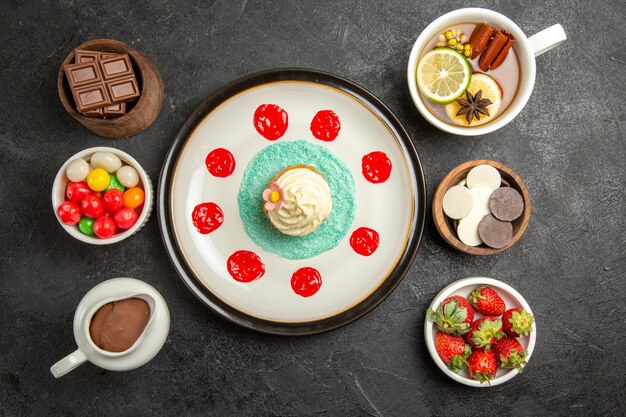 식욕을 돋우는 컵케이크의 차 접시 한 컵, 스타 아니스와 레몬을 넣은 차 한 잔, 검은 테이블에 초콜릿 사탕과 딸기 한 그릇