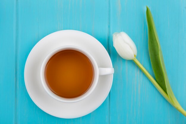 Top view of cup of tea on saucer and flower on blue background