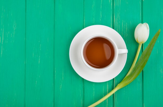 Top view of cup of tea on saucer and flower on green background with copy space