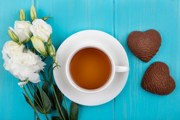 Vista dall'alto della tazza di tè sul piattino e fiori con biscotti a forma di cuore su sfondo blu