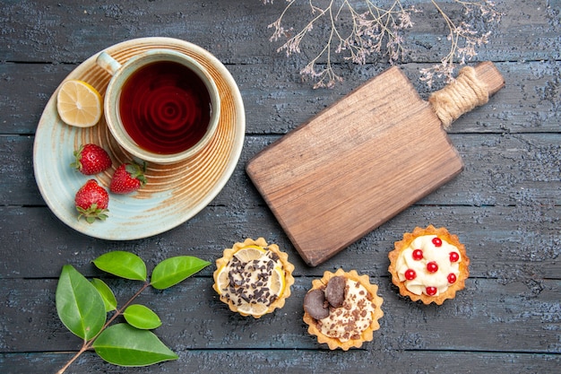 Free photo top view a cup of tea slice of lemon and strawberries on saucer tarts leaves and a chopping board on the dark wooden table