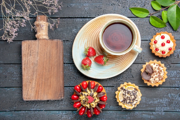 Top view a cup of tea and strawberries on saucer dried oranges tarts leaves berry cake and a chopping board on the dark wooden table