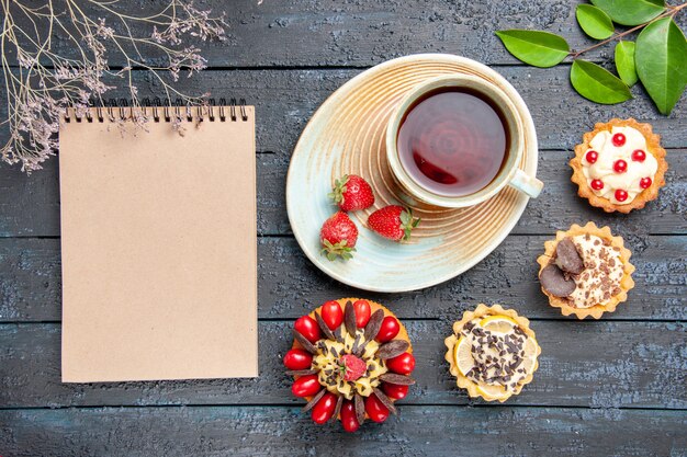 Top view a cup of tea and strawberries on saucer dried oranges tarts leaves berry cake and a notebook on the dark wooden table