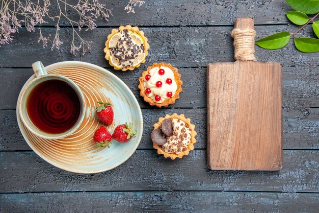 Top view a cup of tea and strawberries on saucer tarts leaves and a chopping board on dark wooden table