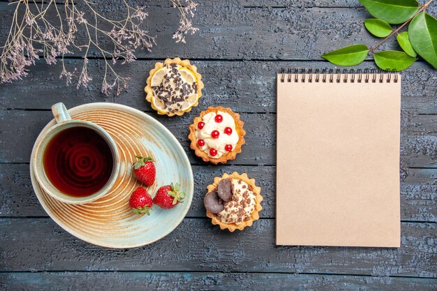 上面図ソーサーのタルトの葉にお茶とイチゴのカップと暗い木製のテーブルにノートブック