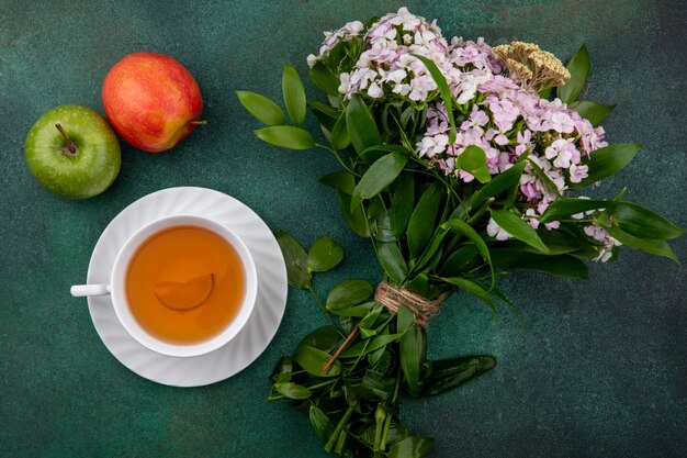 Top view of cup of tea with apples and a bouquet of flowers on a green surface