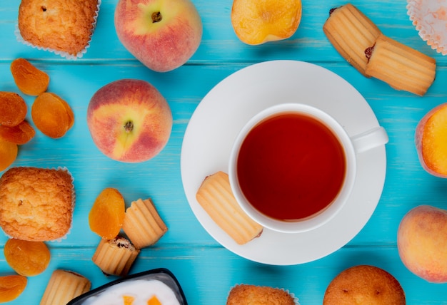 Top view of a cup of tea with biscuits fresh ripe peaches and dried apricots on blue