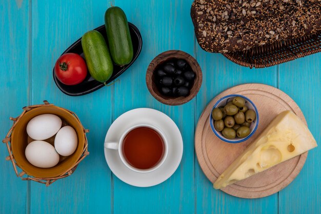 Top view cup of tea with black bread  cheese  cucumbers and tomato and olives with chicken eggs on turquoise background