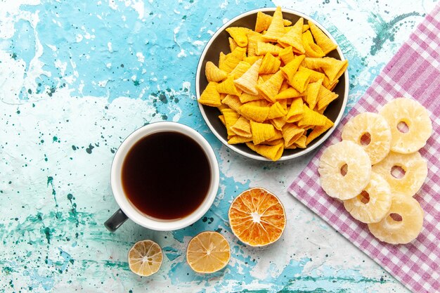 Top view cup of tea with chips and dried pineapple rings on light-blue surface