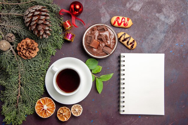 Top view cup of tea with chocolate dessert and notepad on dark space