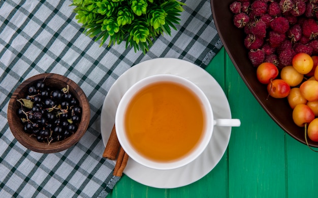 Top view of cup of tea with cinnamon black currants raspberries and white cherries on a checkered green towel