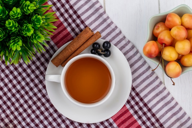 Top view of cup of tea with cinnamon black currants and white cherries on a red checkered towel on a white surface