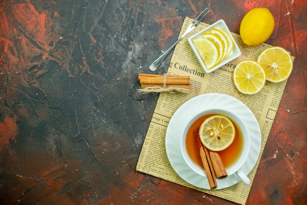 Top view cup of tea with cinnamon lemon slices in small bowl fork on newspaper on dark red background