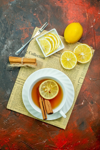 Foto gratuita vista dall'alto tazza di tè con fette di limone alla cannella in una piccola ciotola piccola forchetta sul giornale su sfondo rosso scuro