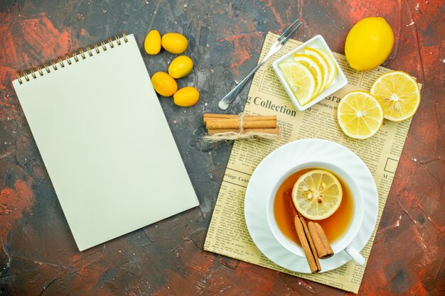 Top view cup of tea with cinnamon lemon slices in small bowl small fork on newspaper notebook cumcuats on dark red background