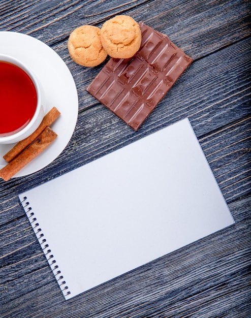 Вид сверху на чашку чая с корицей и альбомом темного шоколада с печеньем на деревенском фоне