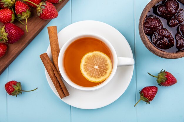 Top view of a cup of tea with cinnamon sticks with strawberry jam on a wooden bowl on a blue background