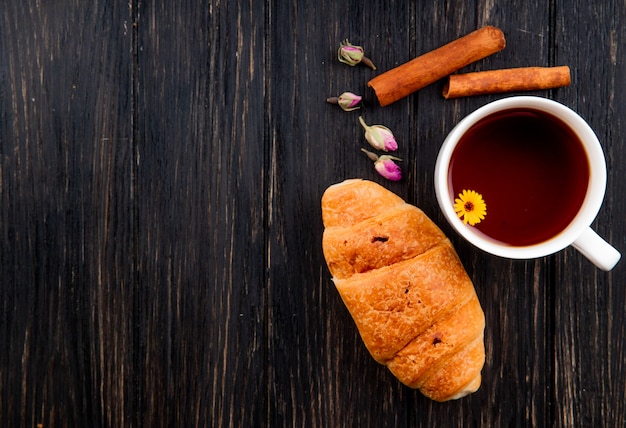 Top view of a cup of tea with croissant and cinnamon sticks on black wood with copy space