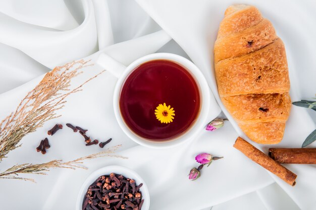 Top view of a cup of tea with croissant and cinnamon sticks and clove spice on white