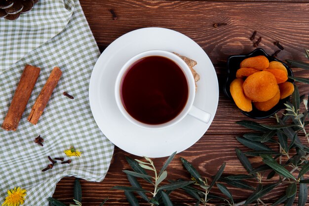 Top view of a cup of tea with dried apricots and cinnamon sticks on wood