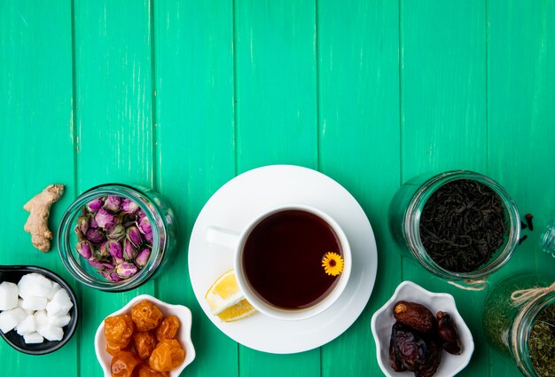 Top view of a cup of tea with dried fruits and dry rose buds with black tea leaves in glass jars on green wood with copy space