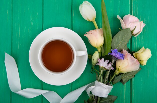 Top view of a cup of tea with flowers like daisy rose and tulip on a green wooden background