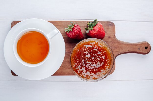 白の木製まな板の上のボウルに新鮮な熟したイチゴとジャムとお茶のカップのトップビュー