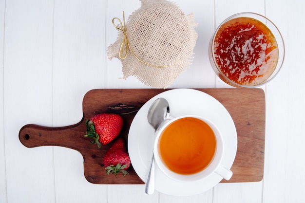 top view of a cup of tea with fresh strawberries on wooden board and strawberry jam in a glass vase on white table
