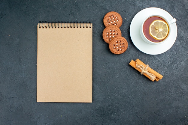 Top view a cup of tea with lemon cinnamon sticks cookies notepad on dark surface