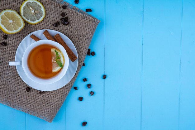 Top view of cup of tea with lemon slice and cinnamon on saucer with lemon slices and chocolate pieces on sackcloth on blue background with copy space