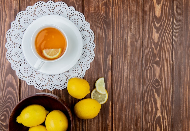 Top view of cup of tea with lemon slice in it on paper doily and lemons on left side and wooden background with copy space