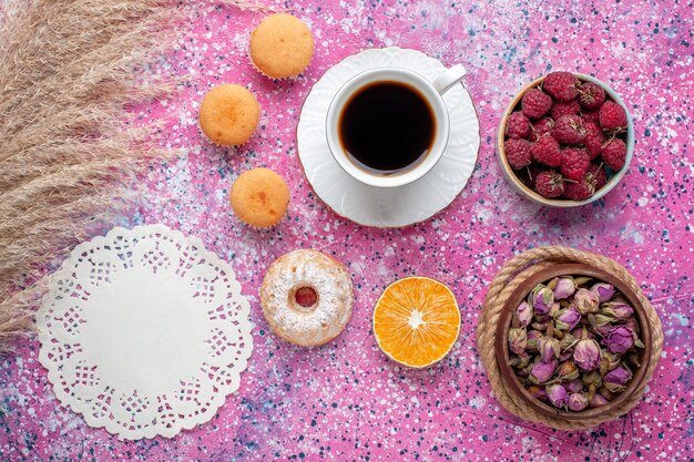 Top view of cup of tea with little cakes and fresh raspberries on pink surface