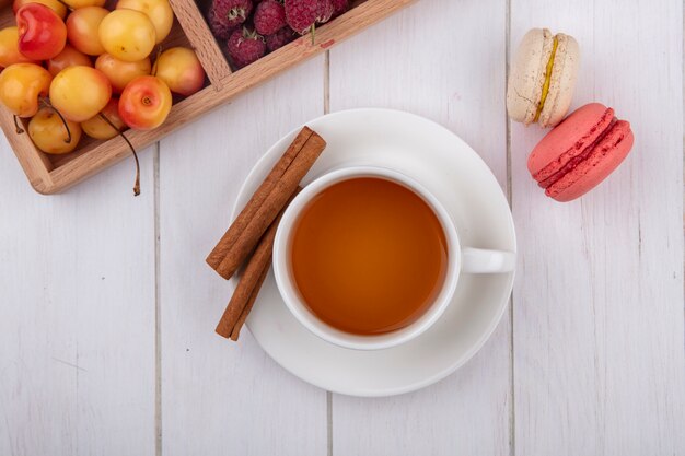 Top view of cup of tea with macarons and cinnamon on a white surface