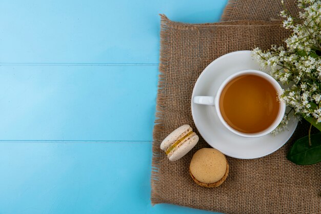 Top view of cup of tea with macarons and flowers on a beige napkin on a blue surface