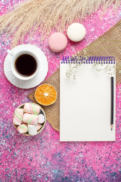 Top view of cup of tea with macarons and marshmallow on pink surface