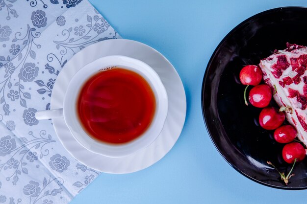 Top view of a cup of tea with a piece of cake decorated with fresh red cherries on black plate on blue