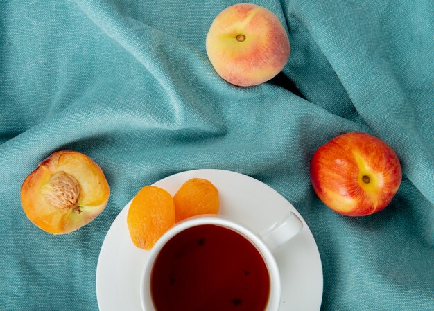 Top view of cup of tea with raisins on teabag and peaches on blue cloth surface