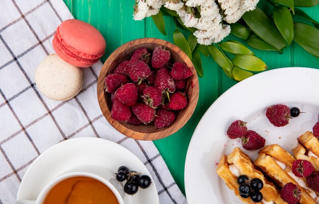 Top view of a cup of tea with raspberries waffles macarons and flowers on a checkered towel