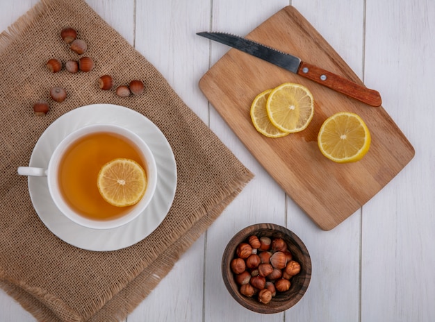 Top view cup of tea with a slice of lemon on a board with a knife and hazelnuts on a gray background